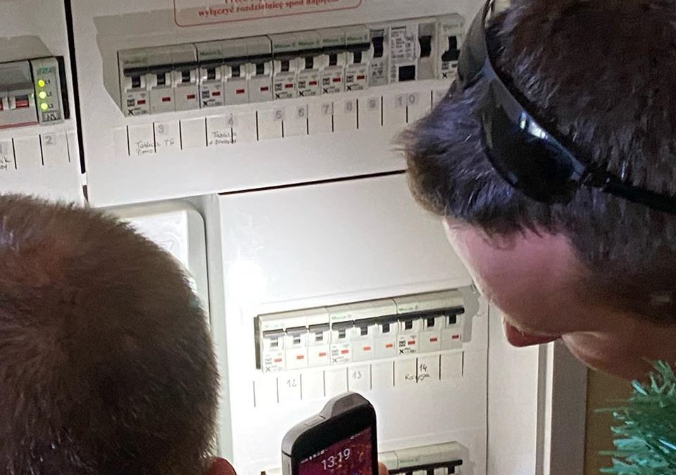 Zdjęcie przedstawia tablicę elektryczną. Patrzy na nią dwóch mężczyzn. Po lewej stronie widać tył głowy jednego z nich, a po prawej lewy profil krótko ostrzyżonego mężczyzny, który świeci na tablicę używając telefonu komórkowego.