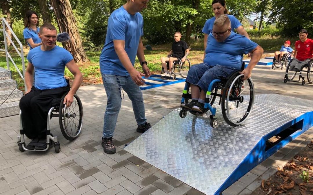 Po pawej stronie - chłopiec ok. 14 lat poruszający się na wózku inwalidzkim zjeżdża z aluminiowej rampy. Asystuje mu wysoki mężczyzna. Po lewej stronie jest dorosły mężczyzna - blondyn w średnim wieku na wózku, który przygląda się chłopcu.